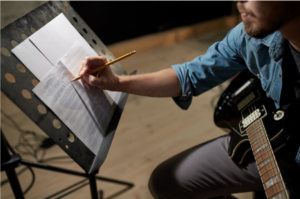 A guy with a guitar in his hand sitting down wrting on sheet music on a music stand.