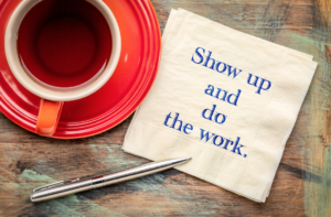 A red plate and a red cup of coffee on a wooden table. with a silver pen and a napkin that reads Show up and do the work.